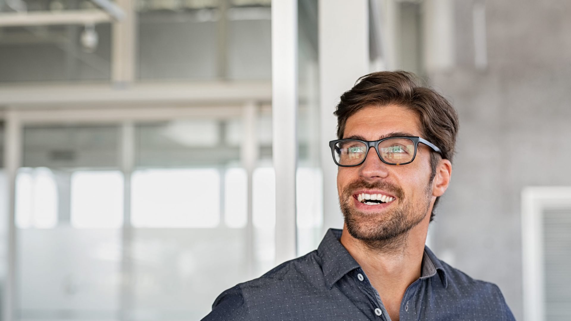 Confident young businessman looking up with big grin. Happy handsome business man wearing spectacles looking away in modern office. Close up face of smiling  guy having idea. Man dreaming of success and pondering new startup.